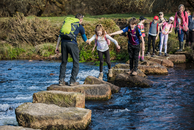 children on a school trip with an outdoor education tutor, crossing a river using boulders