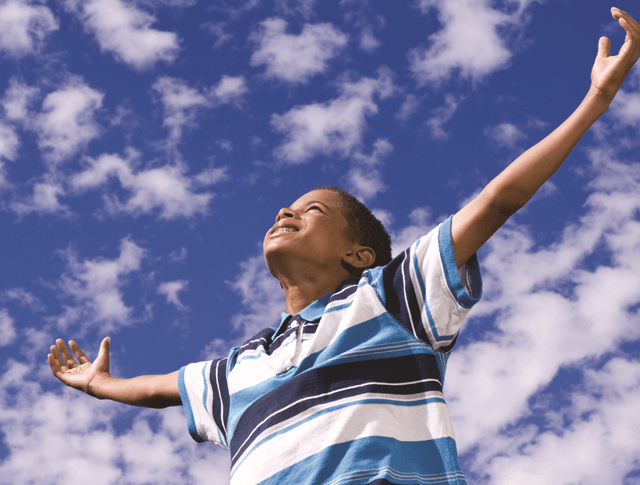 minority ethnic boy with arms outstretched looking up towards a blue and cloudy sky