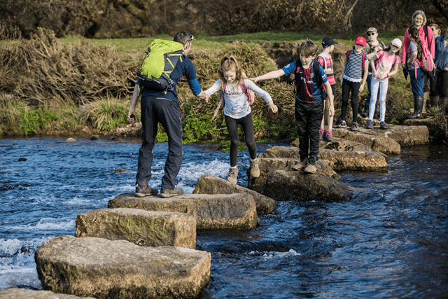 children on a school trip with an outdoor education tutor, crossing a river using boulders